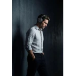 Man in a light shirt with hands in pockets wearing over-ear headphones, standing against a dark concrete wall, looking down.