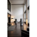 Modern loft living space with tall windows and a high ceiling, featuring two large gray floorstanding speakers and contemporary black seating.